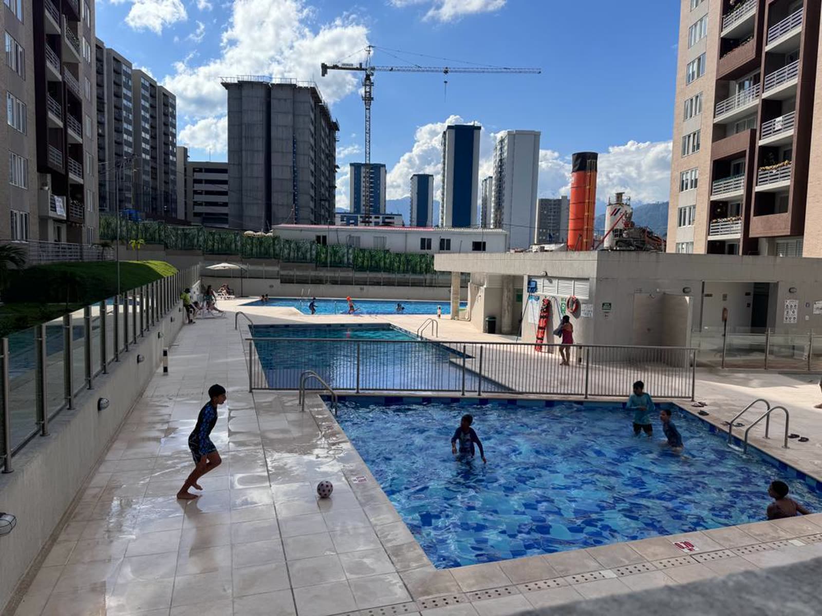 Piscinas del conjunto con niños nadando, torres y grúa al fondo